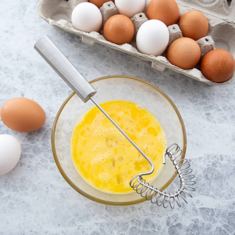 Stainless steel coil whisk with a metal handle resting in a glass bowl of beaten eggs, surrounded by brown and white eggs in a carton on a kitchen countertop, ideal for Kitchen & Food and Cooking and Baking Helpers collections.