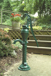 Classic deep well pump head in dark green cast iron with a long lever handle, displayed outdoors near garden plants and steps, part of USA Made water pump collection.