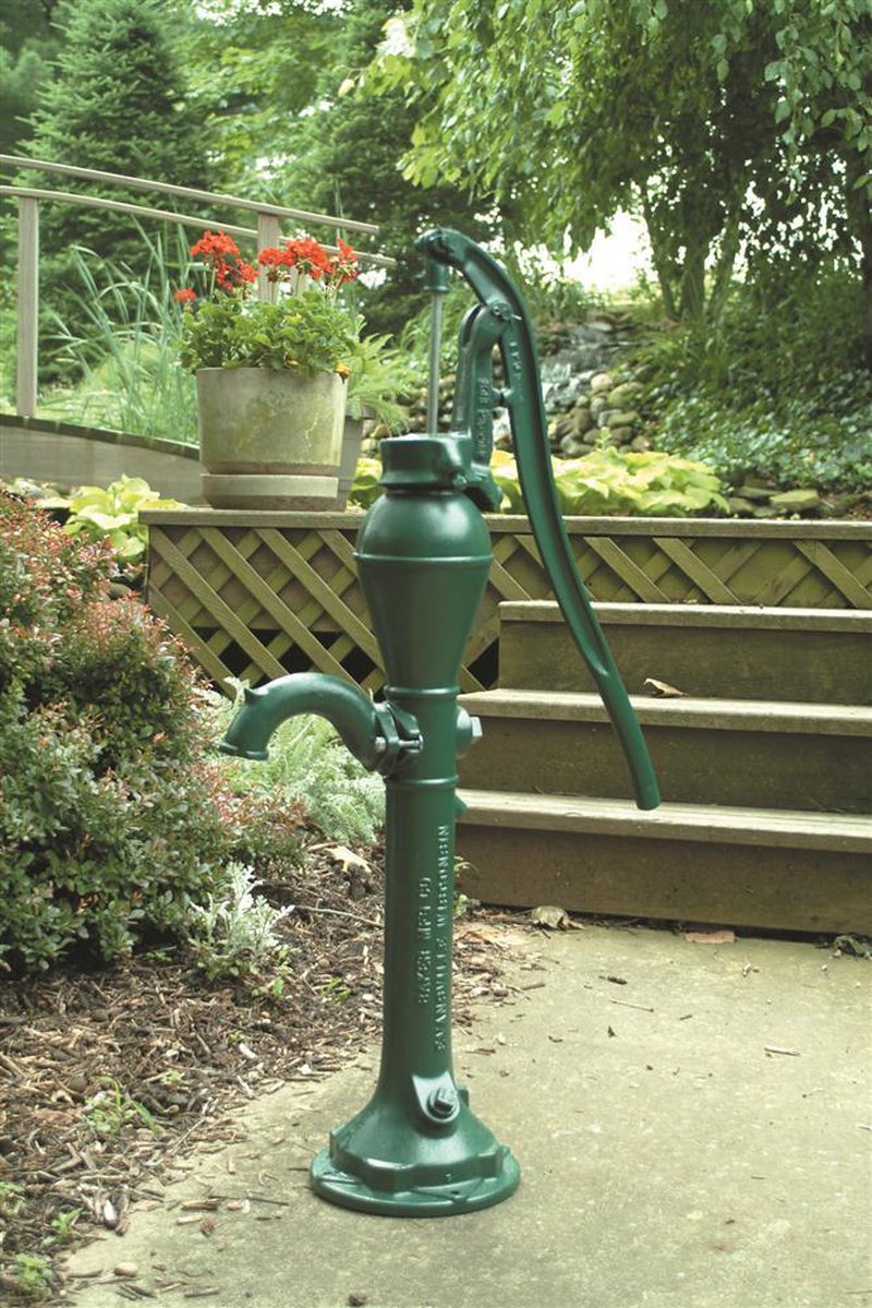 Classic deep well pump head in dark green cast iron with a long lever handle, displayed outdoors near garden plants and steps, part of USA Made water pump collection.