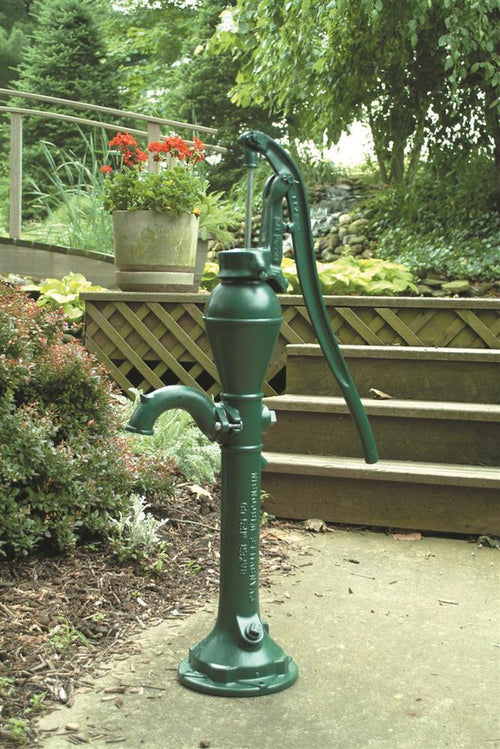 Classic deep well pump head in dark green cast iron with a long lever handle, displayed outdoors near garden plants and steps, part of USA Made water pump collection.
