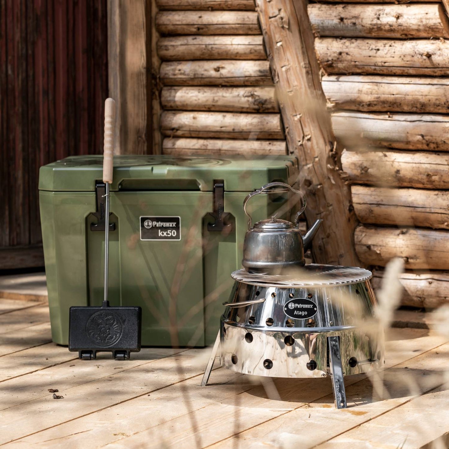 Olive green durable cooler box with heavy-duty latches alongside a stainless steel camping stove and kettle, set on wooden porch with rustic log cabin backdrop, suited for Farm, Garden & Tool, Camping, Best Sellers, and Promo Eligible collections.
