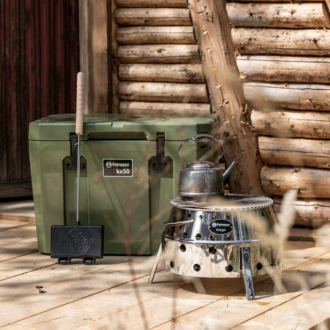 Olive green durable cooler box with heavy-duty latches alongside a stainless steel camping stove and kettle, set on wooden porch with rustic log cabin backdrop, suited for Farm, Garden & Tool, Camping, Best Sellers, and Promo Eligible collections.