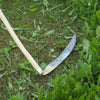 Curved handcrafted scythe blade with a wooden snath resting on green grass, designed for efficient cutting in farm, garden, and lawncare tool collections.