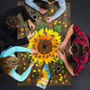 Four people assembling a handcrafted sunflower-shaped 350-piece jigsaw puzzle on a wooden table, surrounded by loose yellow and green puzzle pieces and two mugs, part of Toys, Books & Games and Best Sellers collections.