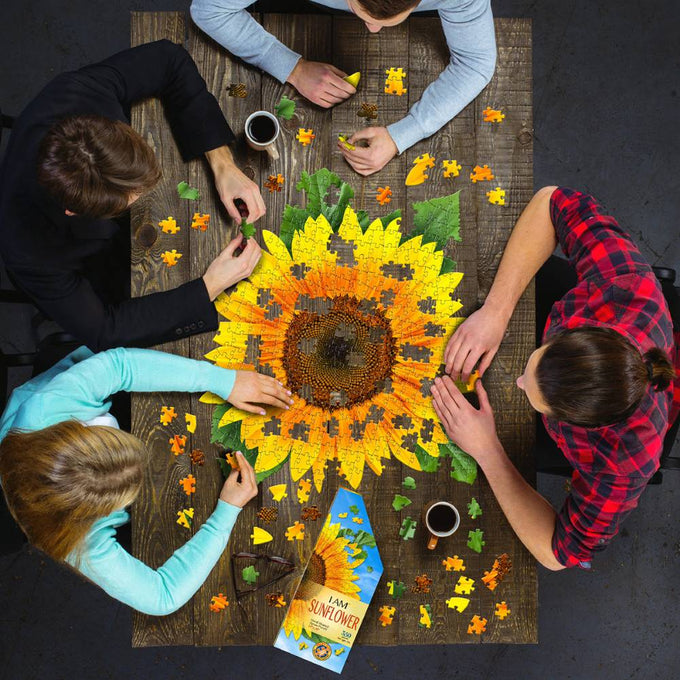 Four people assembling a handcrafted sunflower-shaped 350-piece jigsaw puzzle on a wooden table, surrounded by loose yellow and green puzzle pieces and two mugs, part of Toys, Books & Games and Best Sellers collections.
