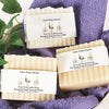Three handcrafted bars of emu oil enriched soap with Dead Sea salt wrapped in rustic labels, displayed on a purple towel with green leaves, highlighting USA made natural wellness products.
