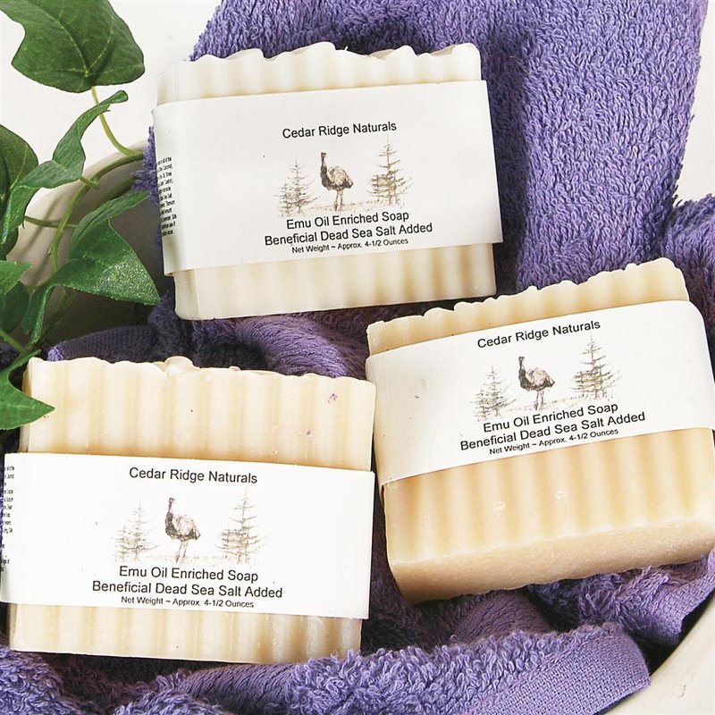 Three handcrafted bars of emu oil enriched soap with Dead Sea salt wrapped in rustic labels, displayed on a purple towel with green leaves, highlighting USA made natural wellness products.