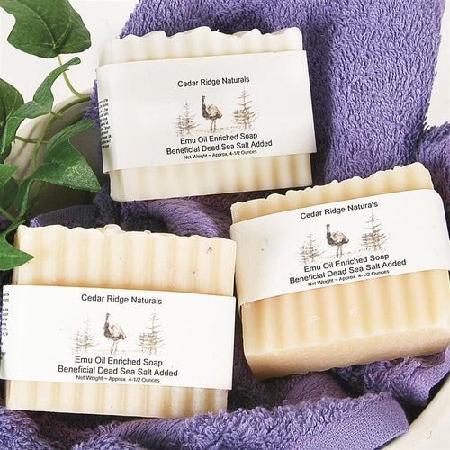 Three handcrafted bars of emu oil enriched soap with Dead Sea salt wrapped in rustic labels, displayed on a purple towel with green leaves, highlighting USA made natural wellness products.