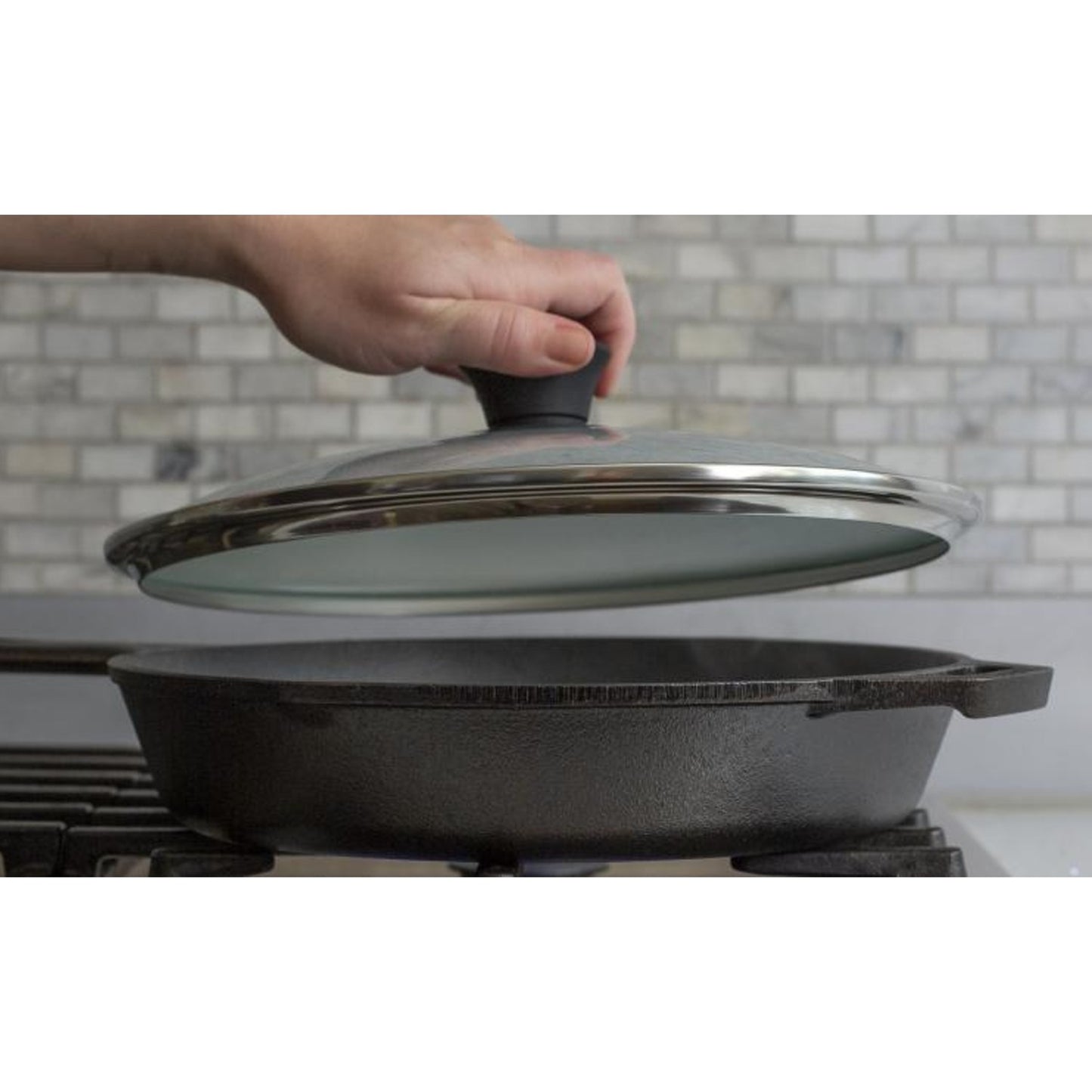 Hand lifting a round tempered glass lid with stainless steel rim and black knob over a cast iron skillet, featured in Kitchen & Food, Cast Iron, Cookware, Pots and Pans, Best Sellers, and Promo Eligible collections.