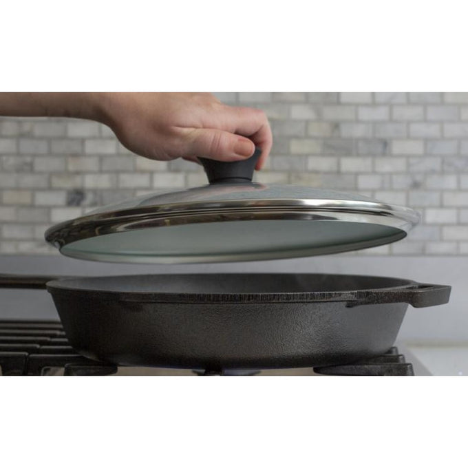 Hand lifting a round tempered glass lid with stainless steel rim and black knob over a cast iron skillet, featured in Kitchen & Food, Cast Iron, Cookware, Pots and Pans, Best Sellers, and Promo Eligible collections.