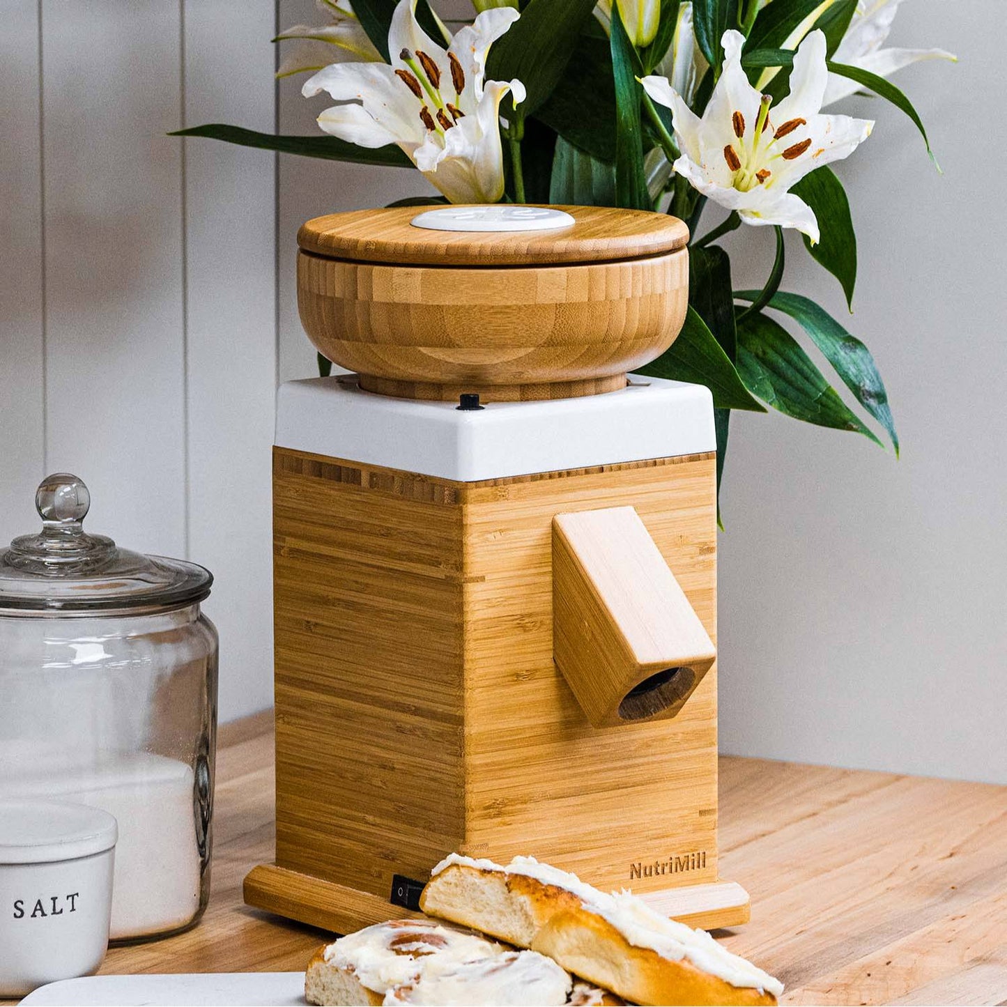 Natural bamboo electric grain mill with a spout for dispensing freshly ground flour, shown on a wooden table with bread and flowers, ideal for Kitchen & Food and Grain Mills collections.