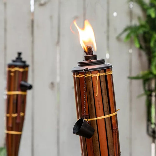 Close-up of a burning bamboo tiki torch with a metal cap and snuffer cup, set against a blurred wooden fence and greenery, fitting Home & Decor, Outdoor Lighting, and Best Sellers collections.