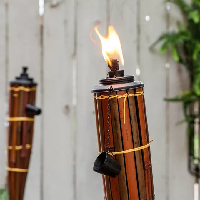 Close-up of a burning bamboo tiki torch with a metal cap and snuffer cup, set against a blurred wooden fence and greenery, fitting Home & Decor, Outdoor Lighting, and Best Sellers collections.