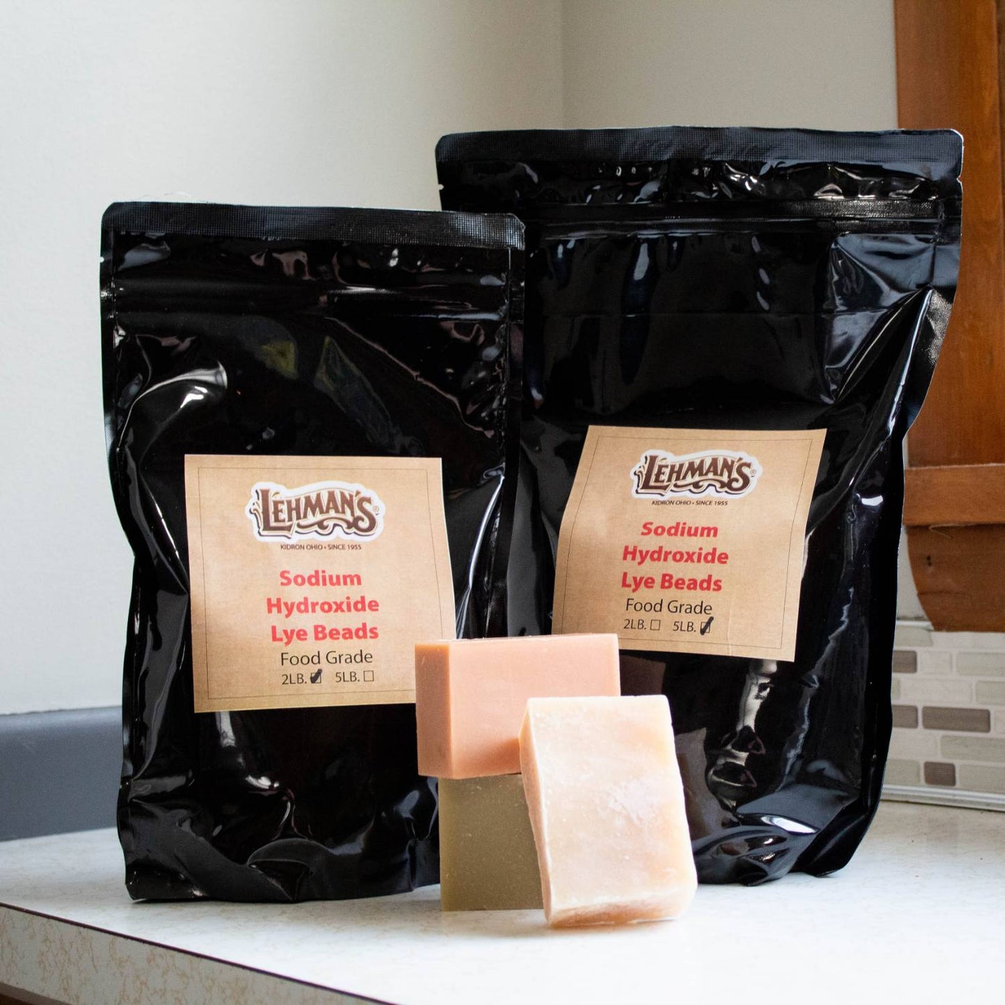 Two resealable black bags of high-grade sodium hydroxide lye beads in 2-lb and 5-lb sizes displayed with three handcrafted soap bars, featured in Health & Wellness, Soaps and Soapmaking, USA Made, and Best Sellers collections.