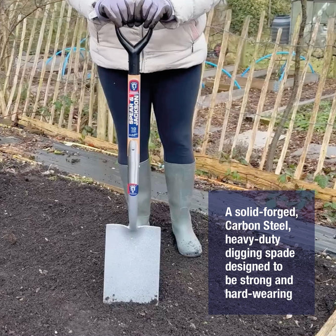 Person wearing gray gloves and green rubber boots holding a heavy-duty, solid-forged carbon steel digging spade on dark garden soil, ideal for Farm, Garden & Tool, Digging and Cultivating, Lawncare, Best Sellers, and Promo Eligible collections.
