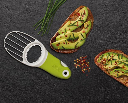 Green-handled 3-in-1 avocado tool with stainless steel blade and slicer resting on dark surface next to two slices of toasted bread topped with sliced avocado, chives, and red pepper flakes, perfect for Kitchen & Food, Food Prep, Choppers and Cutters, and Kitchen Gadgets collections.
