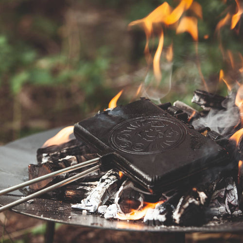 Cast iron waffle iron with long handles cooking over an open campfire, ideal for outdoor dessert making in Farm, Garden & Tool, Camping, Best Sellers, and Promo Eligible collections.