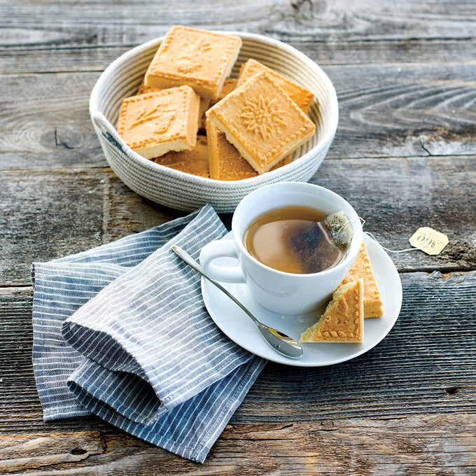 Bowl of golden, square shortbread with decorative patterns, served alongside a white teacup with steeping tea bag, set on rustic wood with striped linen for Kitchen & Food and Baking Supplies collections.