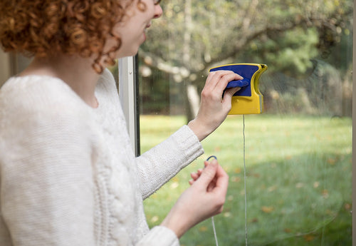 Woman using a handcrafted magnetic window cleaner with yellow and blue plastic housings and a safety rope, shown cleaning windows from inside a home in a Household Cleaning setting.
