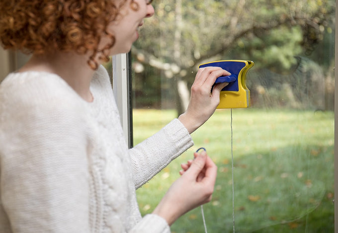Woman using a handcrafted magnetic window cleaner with yellow and blue plastic housings and a safety rope, shown cleaning windows from inside a home in a Household Cleaning setting.