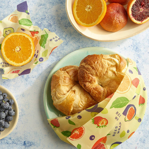 Handmade beeswax food wraps with colorful fruit and leaf patterns covering croissants and a halved orange, eco-friendly and reusable for kitchen storage, part of USA Made, Kitchen & Food collections.