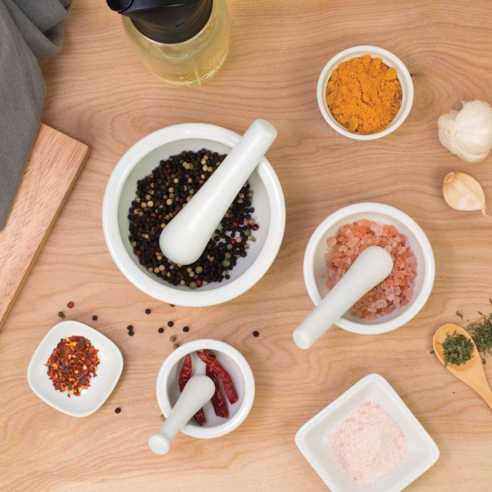 Set of three white porcelain mortars and pestles containing various spices including peppercorns, Himalayan salt, dried chili, and turmeric, styled on a wooden surface with garlic and herbs, featured in Kitchen & Food and Food Prep collections.