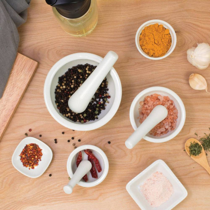 Set of three white porcelain mortars and pestles containing various spices including peppercorns, Himalayan salt, dried chili, and turmeric, styled on a wooden surface with garlic and herbs, featured in Kitchen & Food and Food Prep collections.