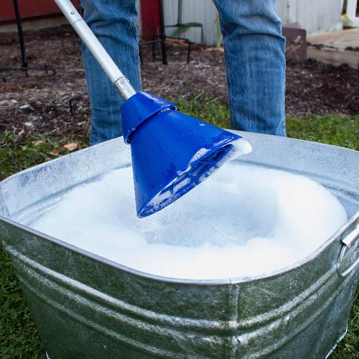 Metal tub filled with soapy water being used with a blue hand-operated collapsible washer plunger on grass outdoors, ideal for Home & Decor and Laundry Supplies collections.