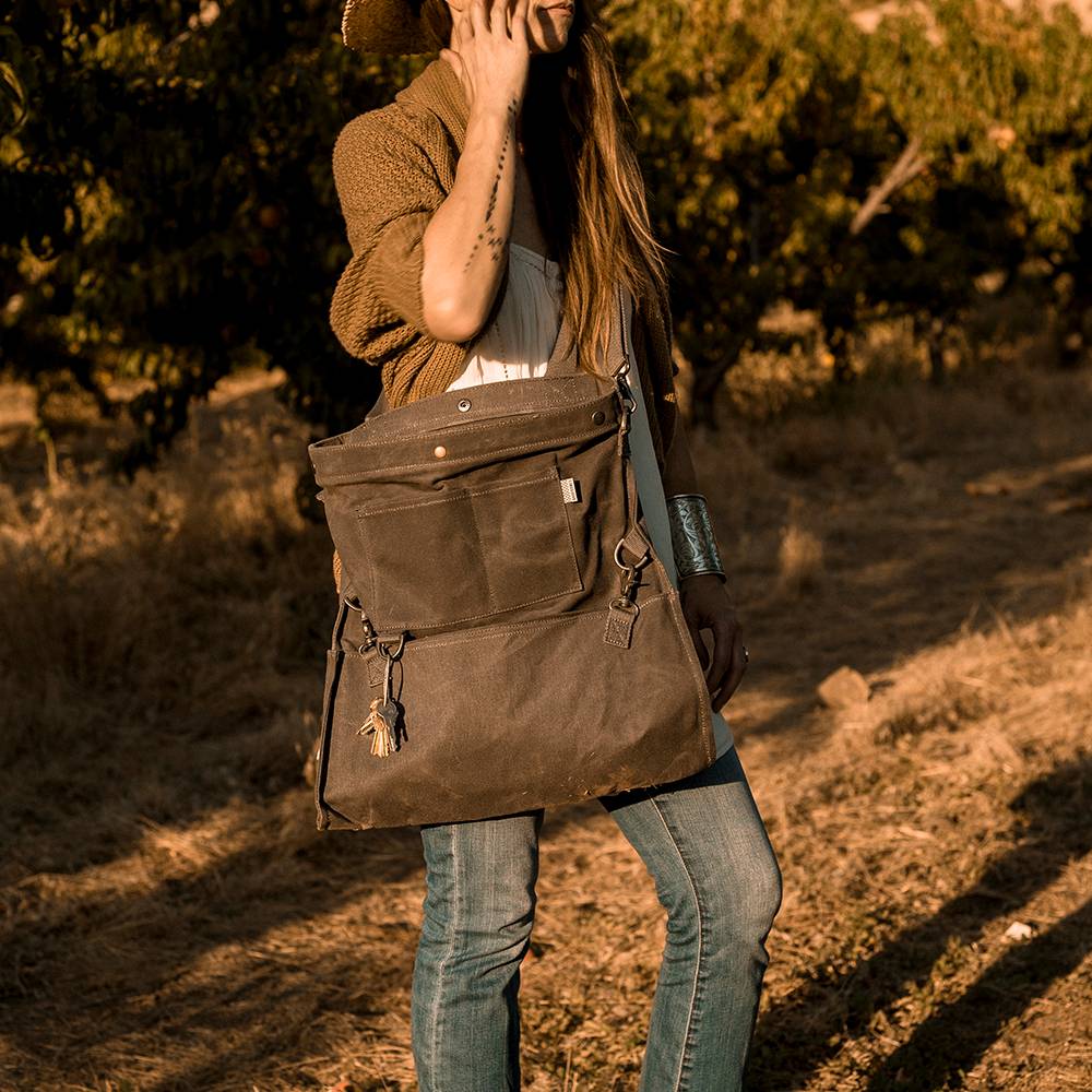 Person standing outdoors in a sunlit orchard wearing a convertible strap bag made of weather-resistant fabric, perfect for farm, garden, and apple harvest supplies.