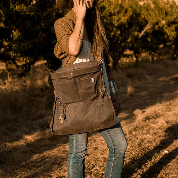 Person standing outdoors in a sunlit orchard wearing a convertible strap bag made of weather-resistant fabric, perfect for farm, garden, and apple harvest supplies.