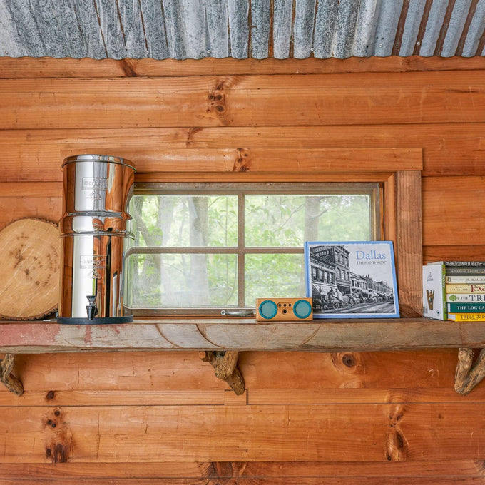 Compact stainless steel gravity water filter with two black purification elements and a spigot, designed for travel and emergency use, displayed on a rustic wooden shelf near a window.