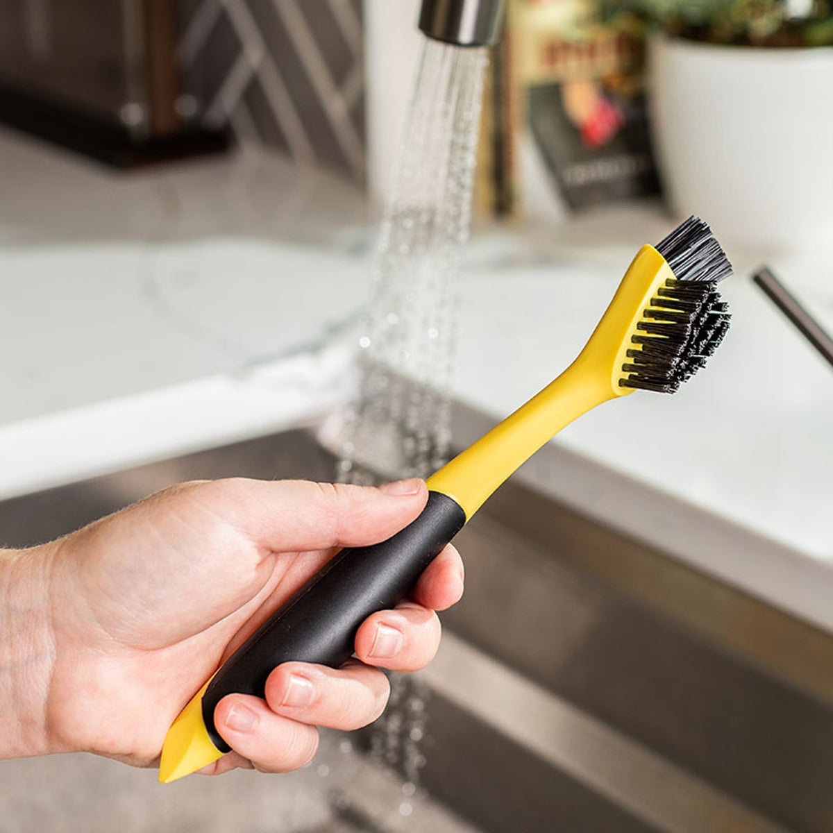 Hand holding a yellow and black corner groove cleaning brush with dual nylon bristle heads under running water, ideal for Kitchen & Food, Cookware, Cast Iron, Best Sellers, and Promo Eligible collections.