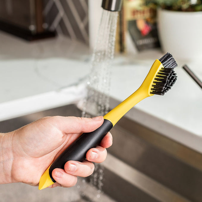 Hand holding a yellow and black corner groove cleaning brush with dual nylon bristle heads under running water, ideal for Kitchen & Food, Cookware, Cast Iron, Best Sellers, and Promo Eligible collections.