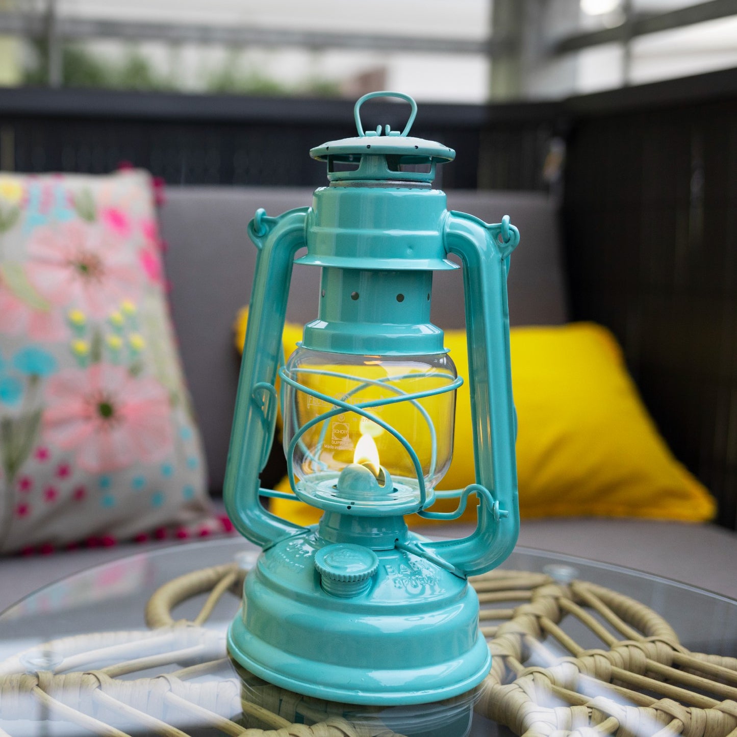 Handcrafted turquoise Feuerhand galvanized oil lantern with glowing thermal heat-resistant SUPRAX globe, placed on glass table in outdoor seating area, ideal for Lamps & Lighting, Camping, and Emergency Supplies collections.