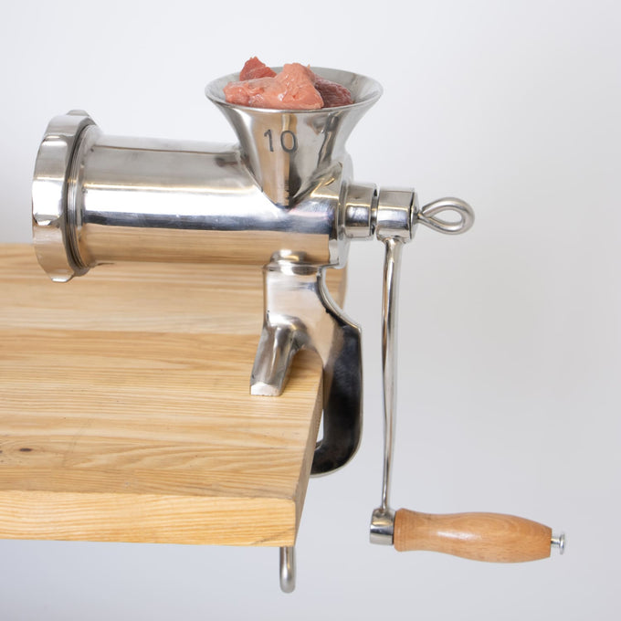 Stainless steel hand-cranked meat grinder with wooden handle clamped to a wooden table, filled with fresh chunks of meat, ideal for Kitchen & Food and Butchering Supplies collections.