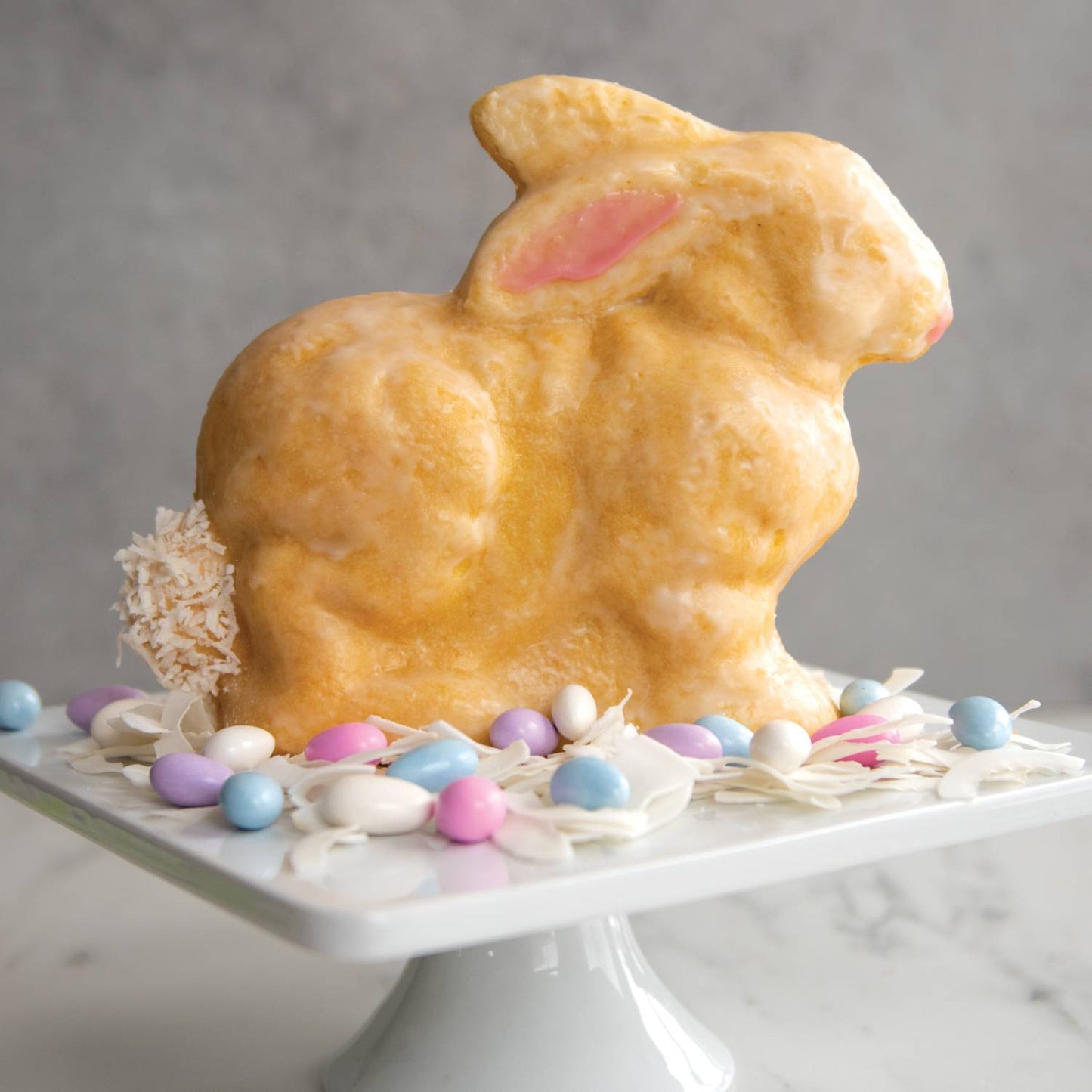 Golden brown 3D bunny-shaped cake with glossy icing and pink accent on ear, displayed on white cake stand surrounded by pastel candy eggs and white flower petals, featured in Kitchen & Food, Cookware, Baking Supplies, USA Made, Best Sellers, and Promo Eligible collections.