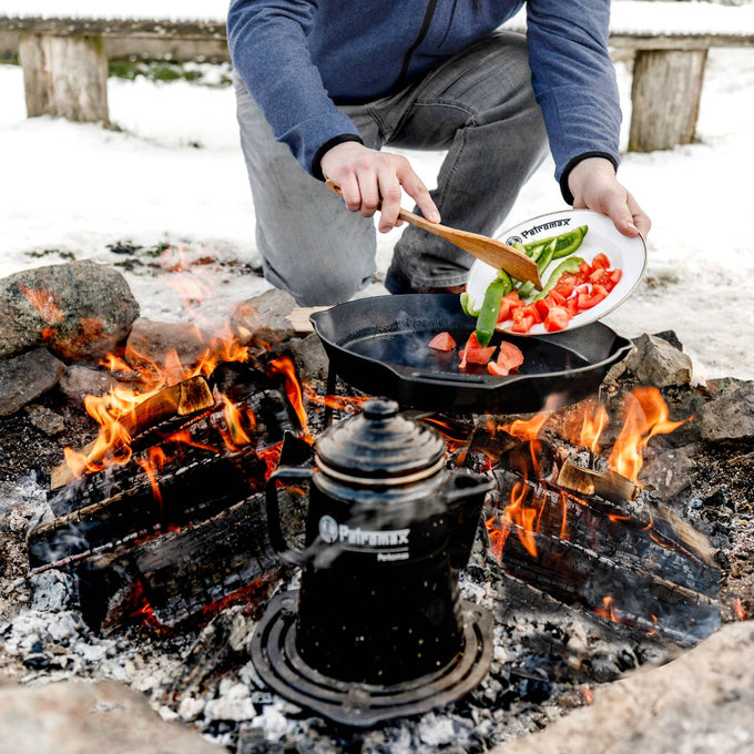 Person adding fresh vegetables from a white enamel Petromax plate into a cast iron pan over an open campfire, surrounded by snow, ideal for Farm, Garden & Tool, Kitchen & Food, Camping, Dinnerware, Best Sellers, and Promo Eligible collections.