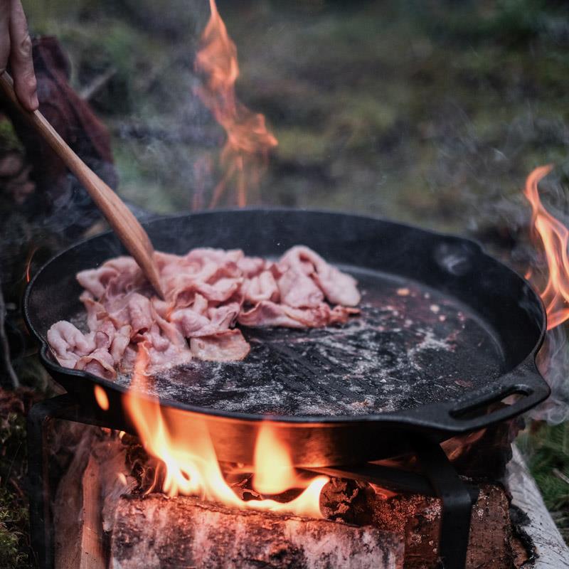Cast iron skillet with long handle cooking strips of meat over an open wood fire, highlighting durable craftsmanship for Farm, Garden & Tool, Kitchen & Food, Camping, Cookware, Cast Iron, Best Sellers, and Promo Eligible collections.