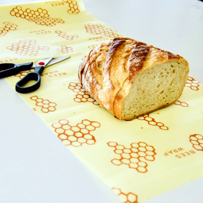 Unrolled beeswax food wrap with honeycomb print beside scissors and a sliced rustic loaf of bread on a white surface, handmade and USA made Kitchen & Food storage item.