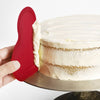 Flexible red silicone scraper with a thin metal core smoothing white frosting on a layered cake, ideal for baking, cookware, and kitchen gadget collections.