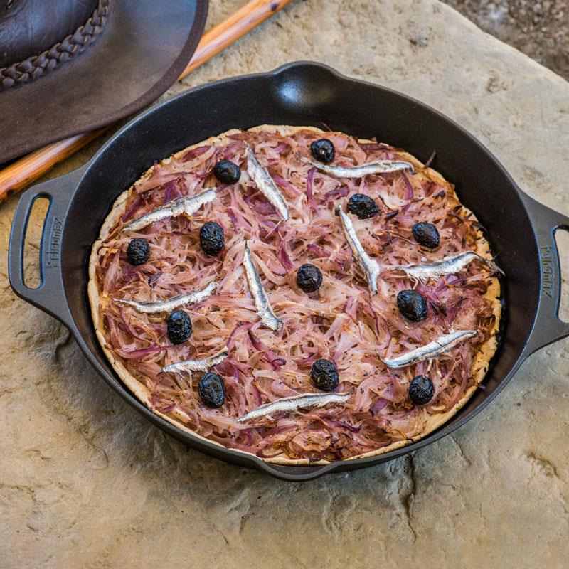 Cast iron skillet with two side handles cooking a rustic pizza topped with anchovies, olives, and caramelized onions, ideal for farm, kitchen, camping, and cookware collections.