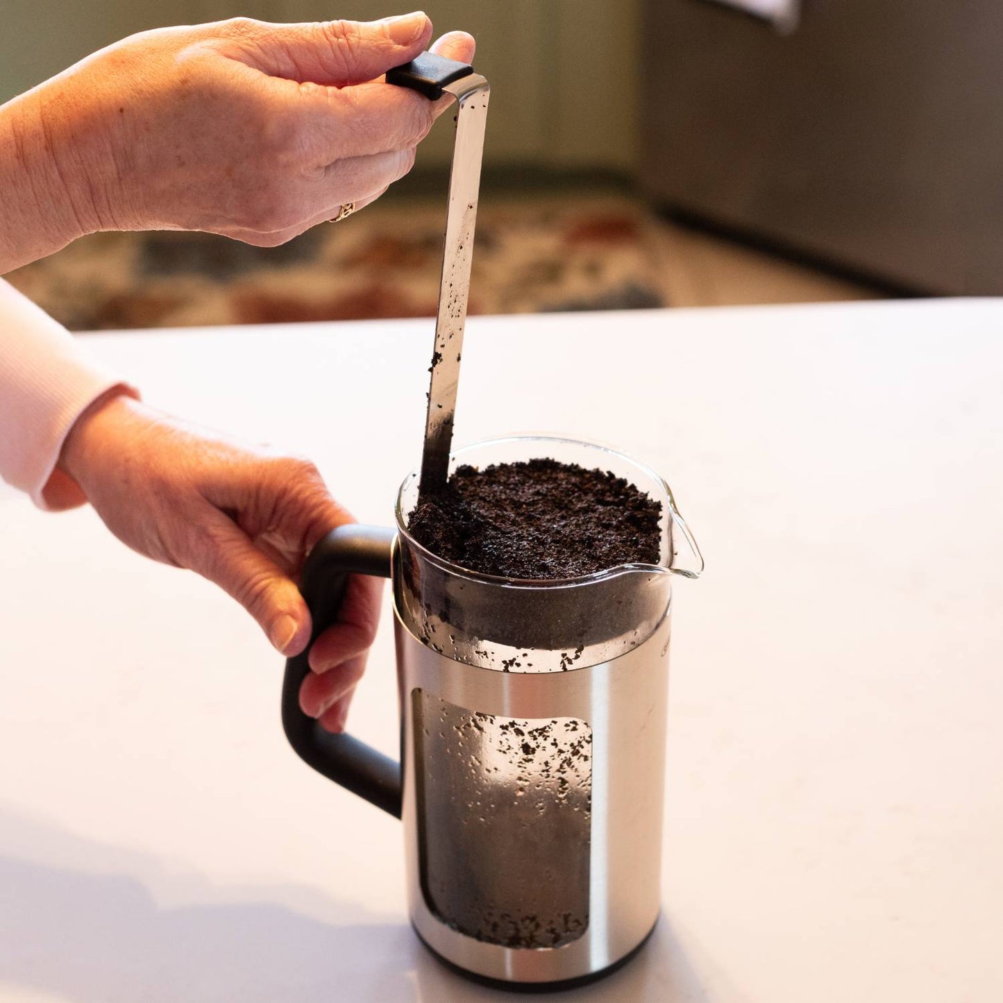 Hands using the GroundsLifter tool to lift coffee grounds from a stainless steel and glass French press coffee maker, suitable for Kitchen & Food and Brewing Coffee and Tea collections.