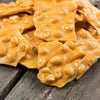 Close-up of thin, crispy peanut brittle pieces with a rich golden-butter color and whole peanuts scattered throughout, handcrafted and made in USA, ideal for Kitchen & Food and Snacks collections.