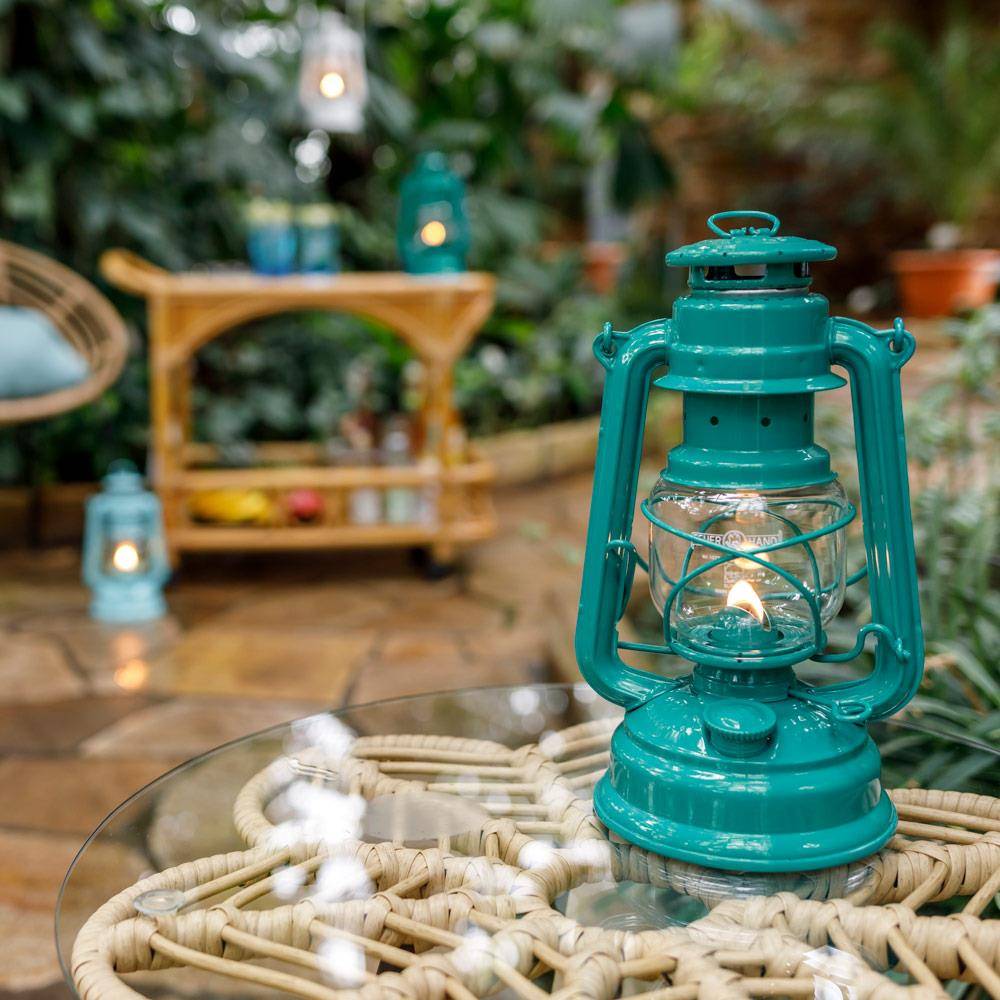 Handcrafted teal galvanized oil lantern with thermal heat-resistant globe softly glowing on a glass table with woven details, surrounded by a lush garden setting, perfect for Camping and Lamps & Lighting collections.