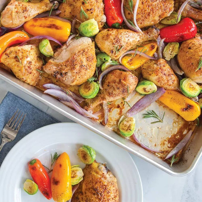 High-sided aluminum pan filled with roasted seasoned chicken thighs, mini bell peppers, Brussels sprouts, and red onion slices, shown with a plated serving, ideal for Kitchen & Food and Cookware collections.