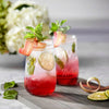 Two handcrafted Ball flute pint jars filled with a red beverage, garnished with mint, lime slices, and rhubarb ribbons, placed on a gray wooden board in a kitchen setting.