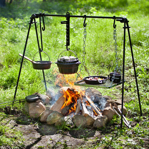 Black-coated steel fire bridge suspended over a campfire with hanging cast iron pots and grill grate cooking food, suitable for Farm, Garden & Tool, Stoves, Camping, Grilling, and Best Sellers collections.