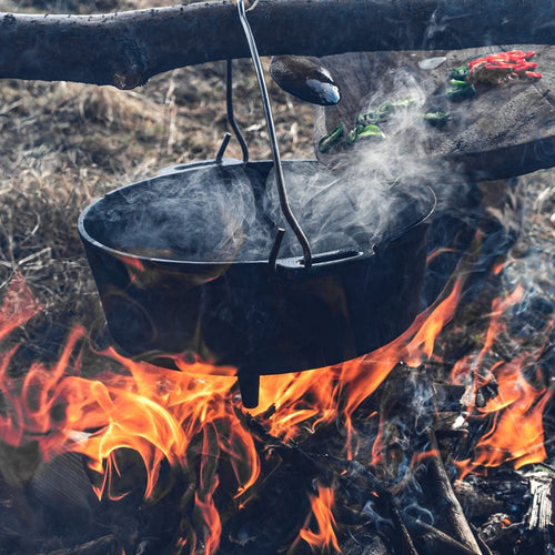 Black cast iron Dutch oven with legs suspended over an open campfire, steaming as it cooks outdoors, ideal for Kitchen & Food, Cast Iron, Cookware, Best Sellers, and Promo Eligible collections.