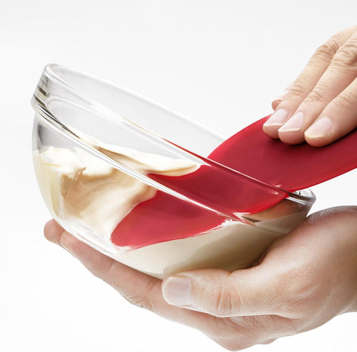 Red flexible scraper with a thin metal core, scraping batter from a clear glass bowl, designed for Kitchen & Food, Cookware, and Baking Supplies collections.