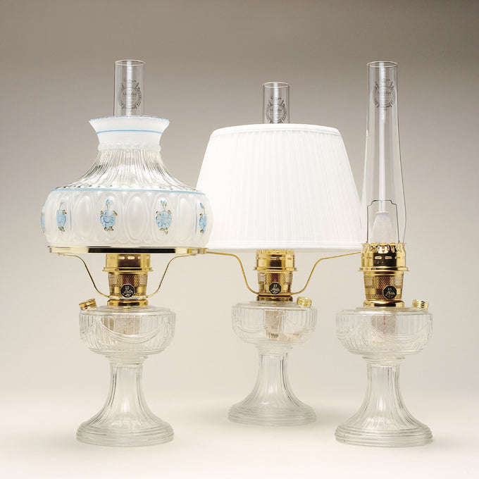 Handcrafted clear glass oil lamps featuring polished brass burners with tall chimneys, including one with a decorative painted shade, part of Lamps & Lighting, Oil Lamps, Table Lamps, Best Sellers, and Promo Eligible collections.
